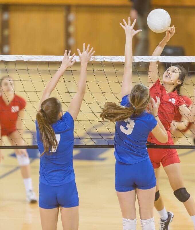 Girl spiking a volleyball over a tall net while two girls jump to block it